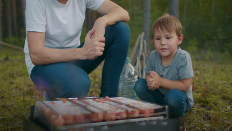 adult-man-and-little-boy-are-sitting-near-chargrill-with-meat-and-sausages-at-nature-picnic-of-family-in-forest-father-and-son